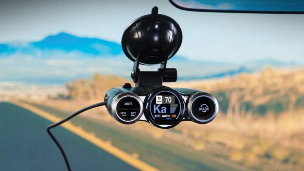 A windshield-mounted black radar detector and a dash cam with a mountain landscape in the background.